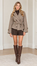 Front view of model wearing brown houndstooth plaid double-breasted belted jacket with brown shorts and tall boots