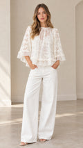 Front full-body view of model wearing white floral lace tiered ruffle blouse with flared sleeves and white wide-leg trousers