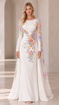 Front view of woman wearing white long-sleeve evening gown with colorful 3D floral embroidery and cape sleeve