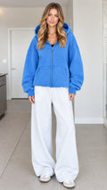 Front full-length view of a woman wearing an oversized blue zip-up fleece hoodie with white sweatpants