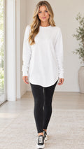 Front full view of a woman wearing a white crewneck long sleeve curved hem tunic top with black leggings
