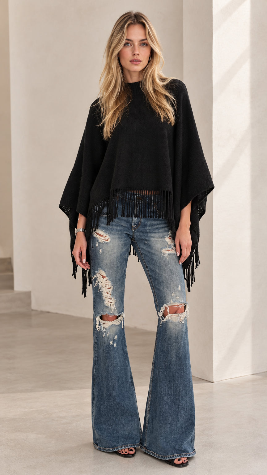 Front view of model wearing black fringe knit poncho sweater and distressed flare jeans