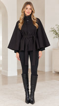 Front view of woman wearing black high-neck belted cape coat with tall black boots