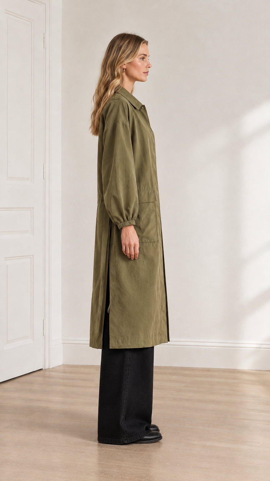 Side profile view of a model in an olive green long duster coat highlighting the high side hem slit