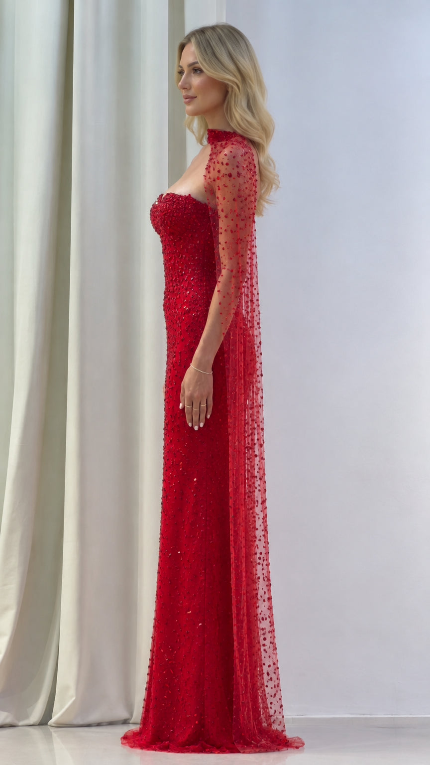 Side profile of model in a red beaded formal maxi dress with flowing sheer cape