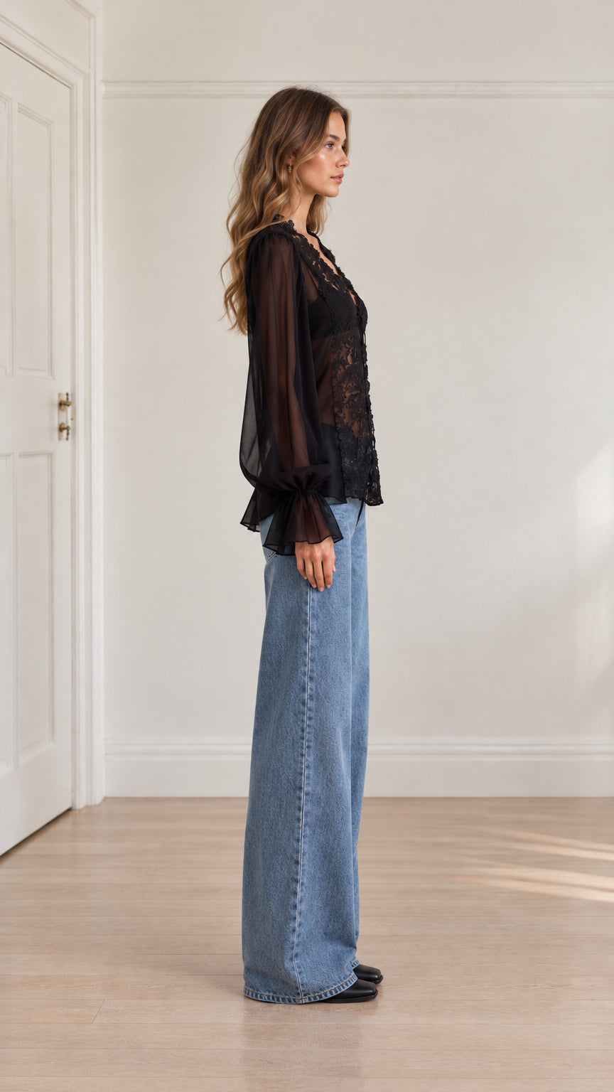 Side angle view of model wearing black sheer lace blouse and denim trousers