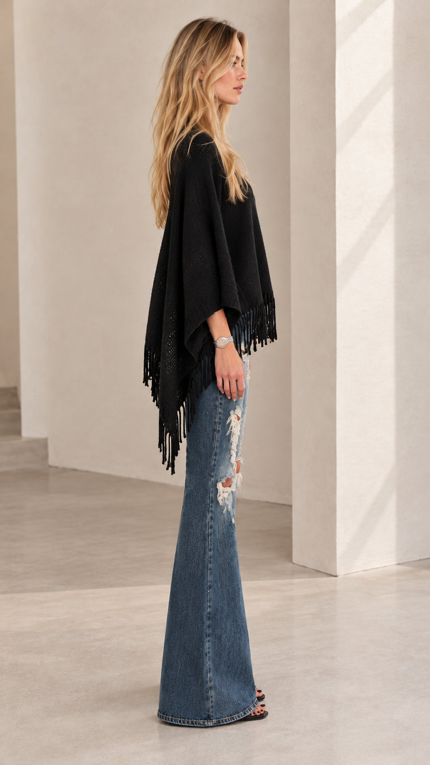 Side profile of model wearing black fringe poncho top with wide leg flare denim pants