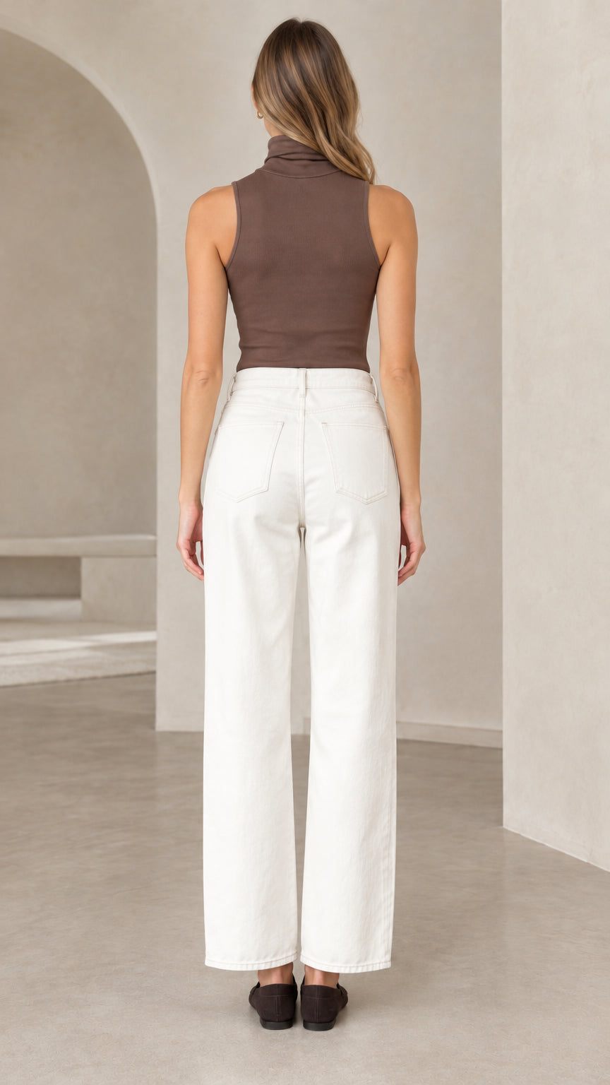 Back view of woman wearing fitted brown sleeveless mock neck knit top with white trousers