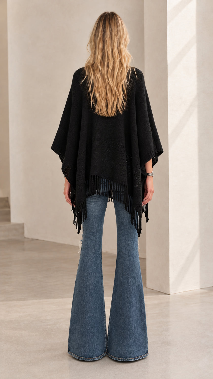 Back view of model wearing black cape poncho sweater and distressed flare jeans