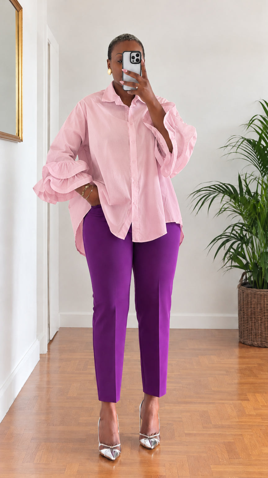 Full-length view of woman wearing light pink tiered ruffle sleeve button-down tunic blouse with purple trousers