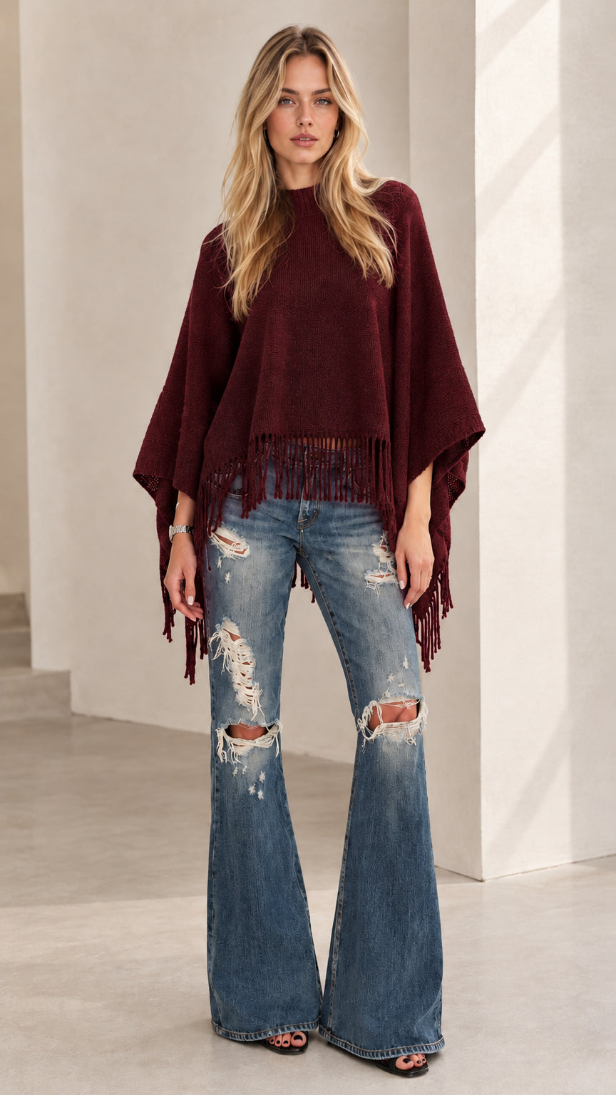 Model wearing burgundy red fringe poncho sweater paired with ripped flare jeans
